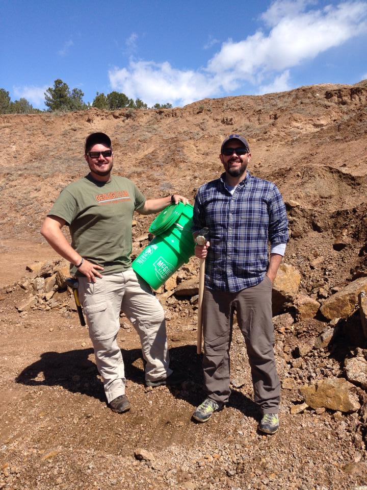 Two geologists standing in a quarry with sample buckets and a big sledge hammer